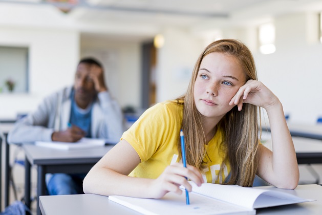 Thoughtful teenage girl leaning on desk. Female student is sitting with pencil and book in classroom. She is against male friend at school.